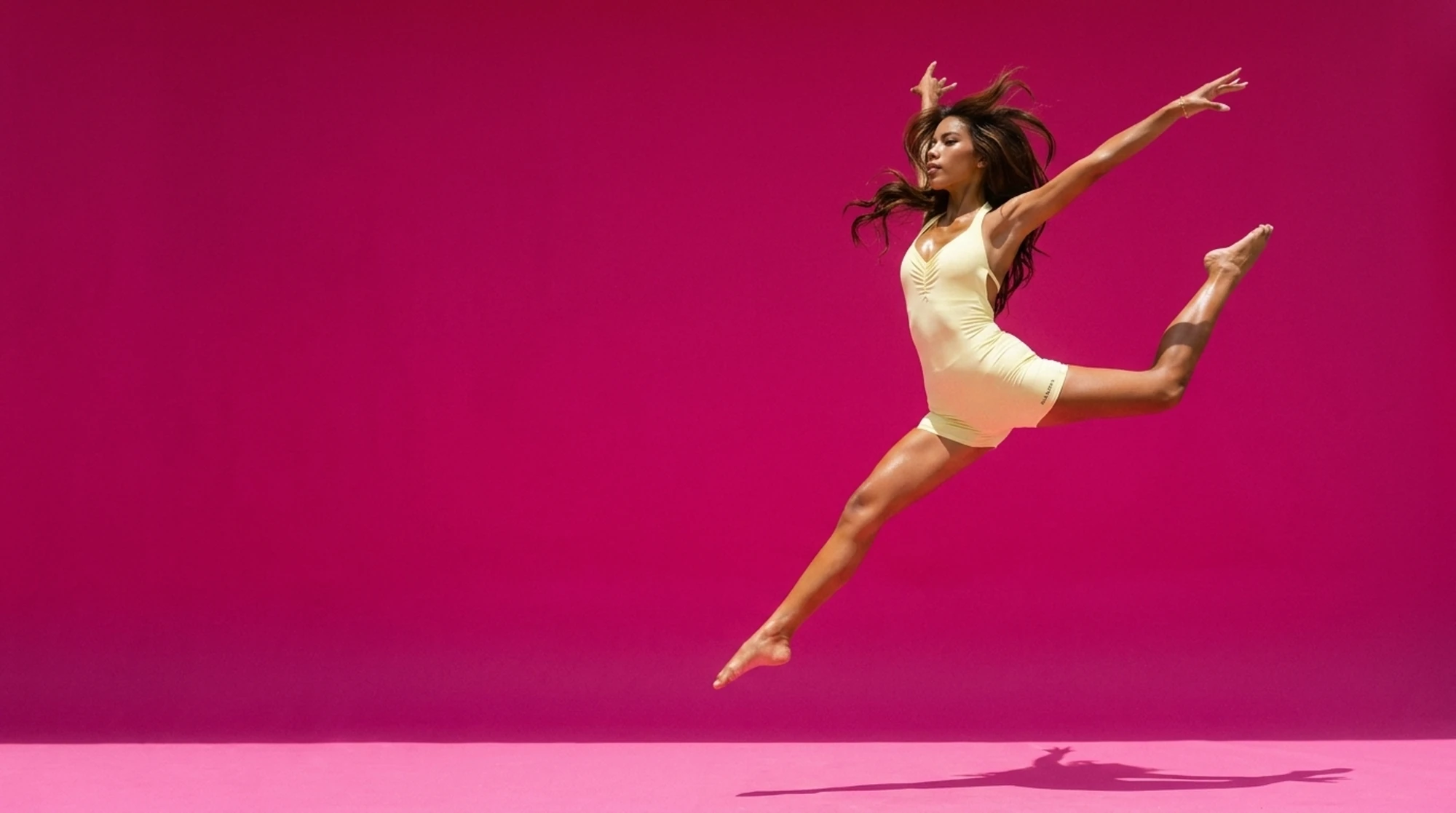 AURANOVA founder mid-leap in the buttercup Nova Jumpsuit on a hot-pink studio sweep